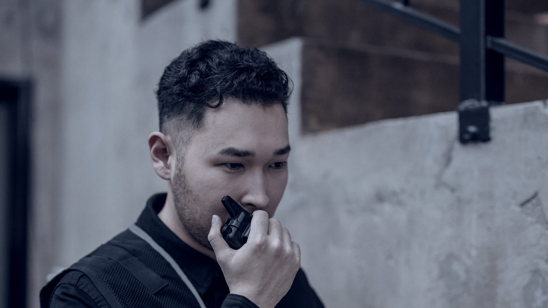 A security officer in a dark jacket and vest speaking into a hand-held radio, seen in profile with his face turned down and away from the camera, against a bare concrete stairwell wall.