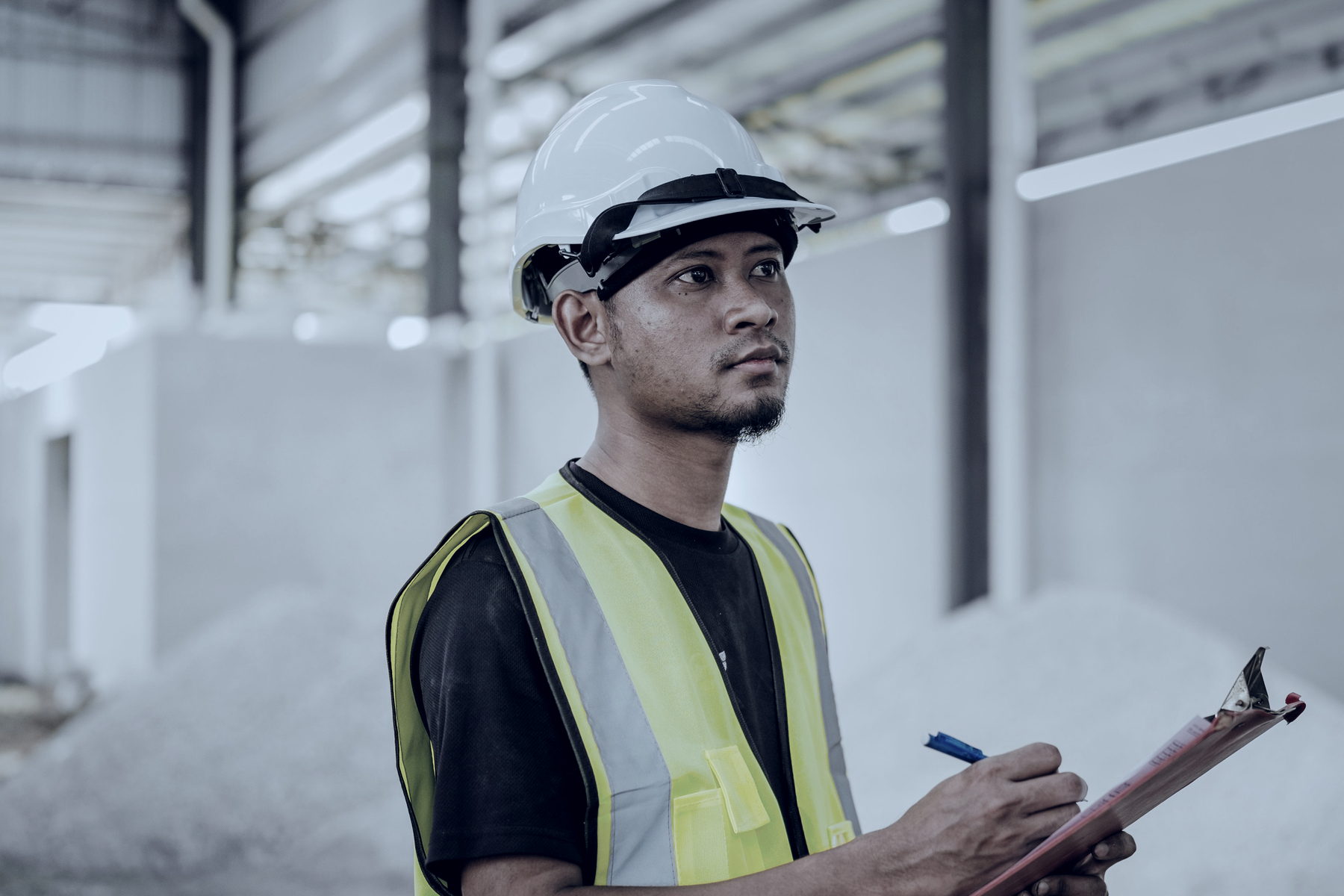 A consultant in a yellow hard hat and hi-vis vest, seen in profile, checking the top hinge of an open door with a pen in one hand and a clipboard in the other.