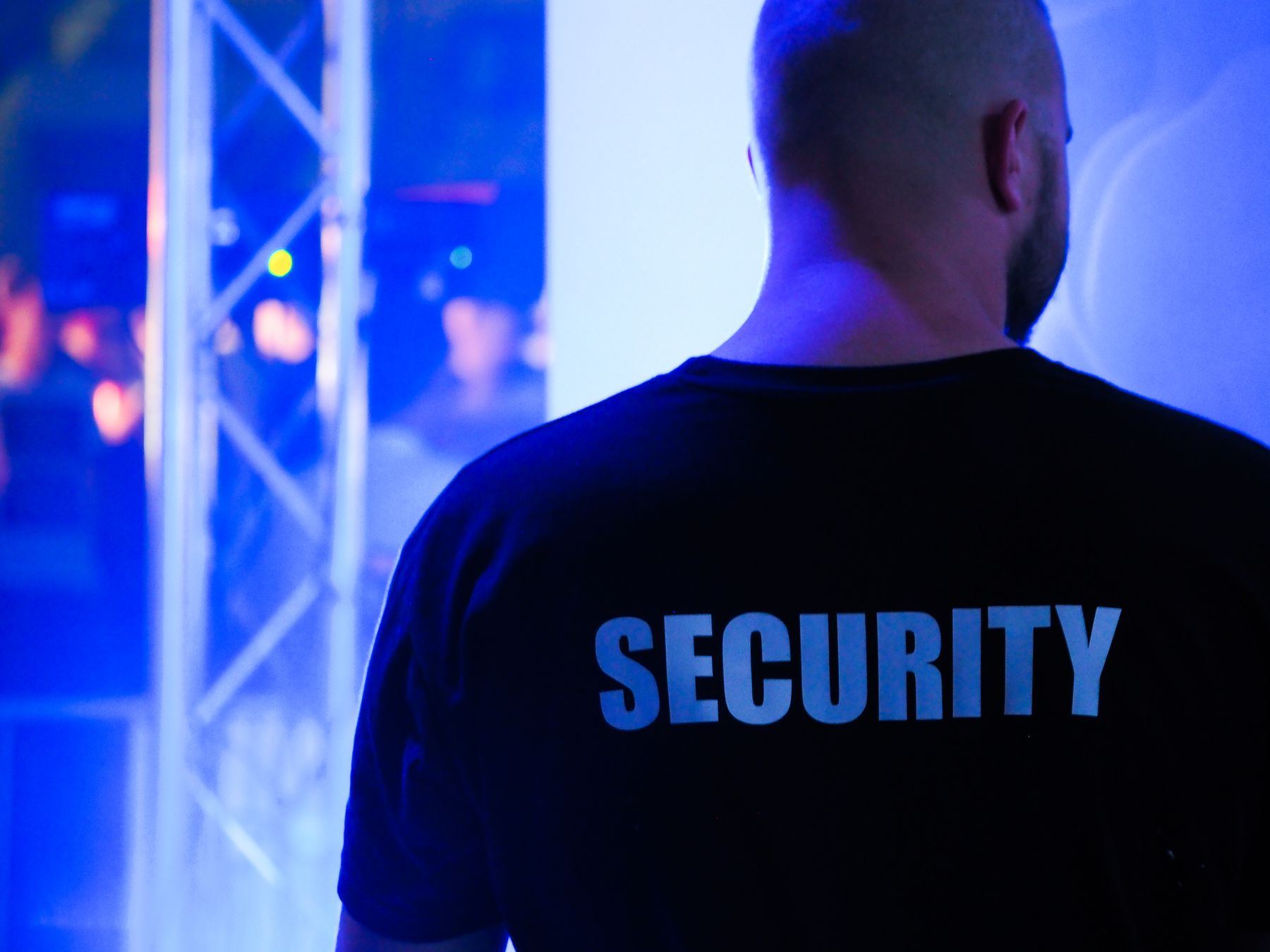 A security officer seen from behind, SECURITY printed across the back of his dark shirt, standing beside a lighting truss under blue venue lighting.