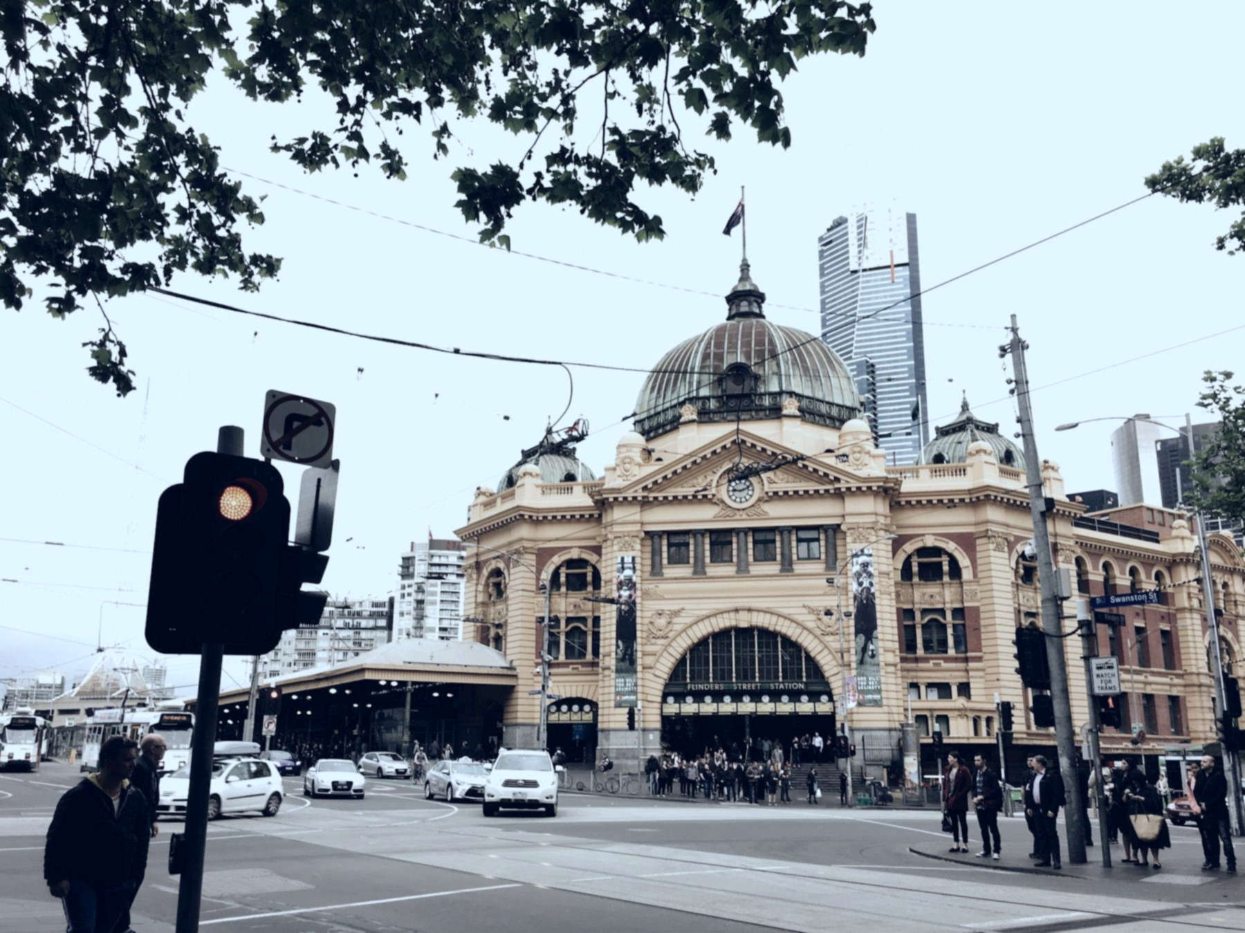 Flinders Street Station on a grey day, with trams, cars and pedestrians at the Swanston Street intersection in Melbourne&rsquo;s CBD.