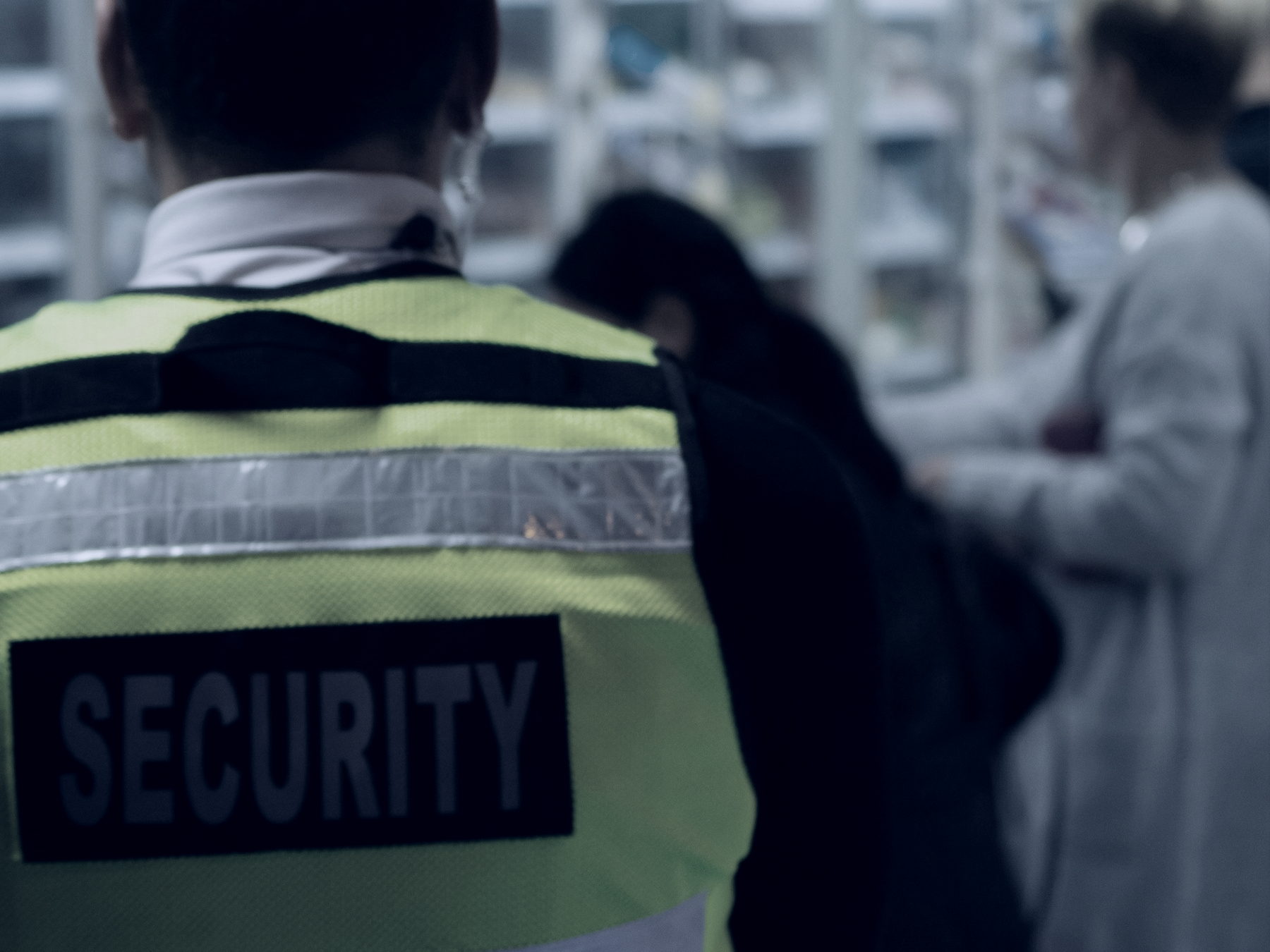 A loss-prevention officer in a hi-vis vest lettered SECURITY, seen from close behind, watching two shoppers at the shelves of a store.