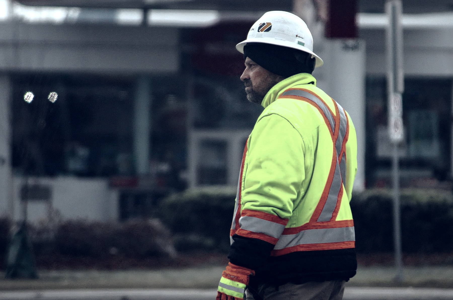 A single worker in a lime hi-vis jacket with orange and silver reflective banding, a white hard hat and heavy gloves, standing in sharp profile at dusk with his face turned away from the camera and a commercial building frontage out of focus behind him.