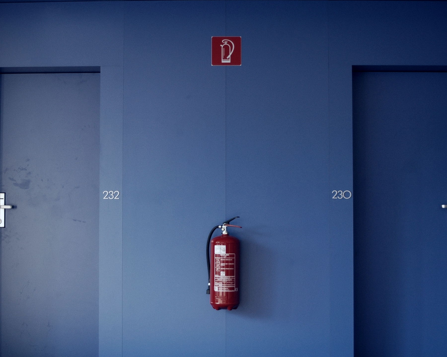 A single red portable fire extinguisher wall-mounted in a blue-panelled commercial corridor, its white service label intact on the cylinder, below the red fire-equipment location sign and between two office doors.