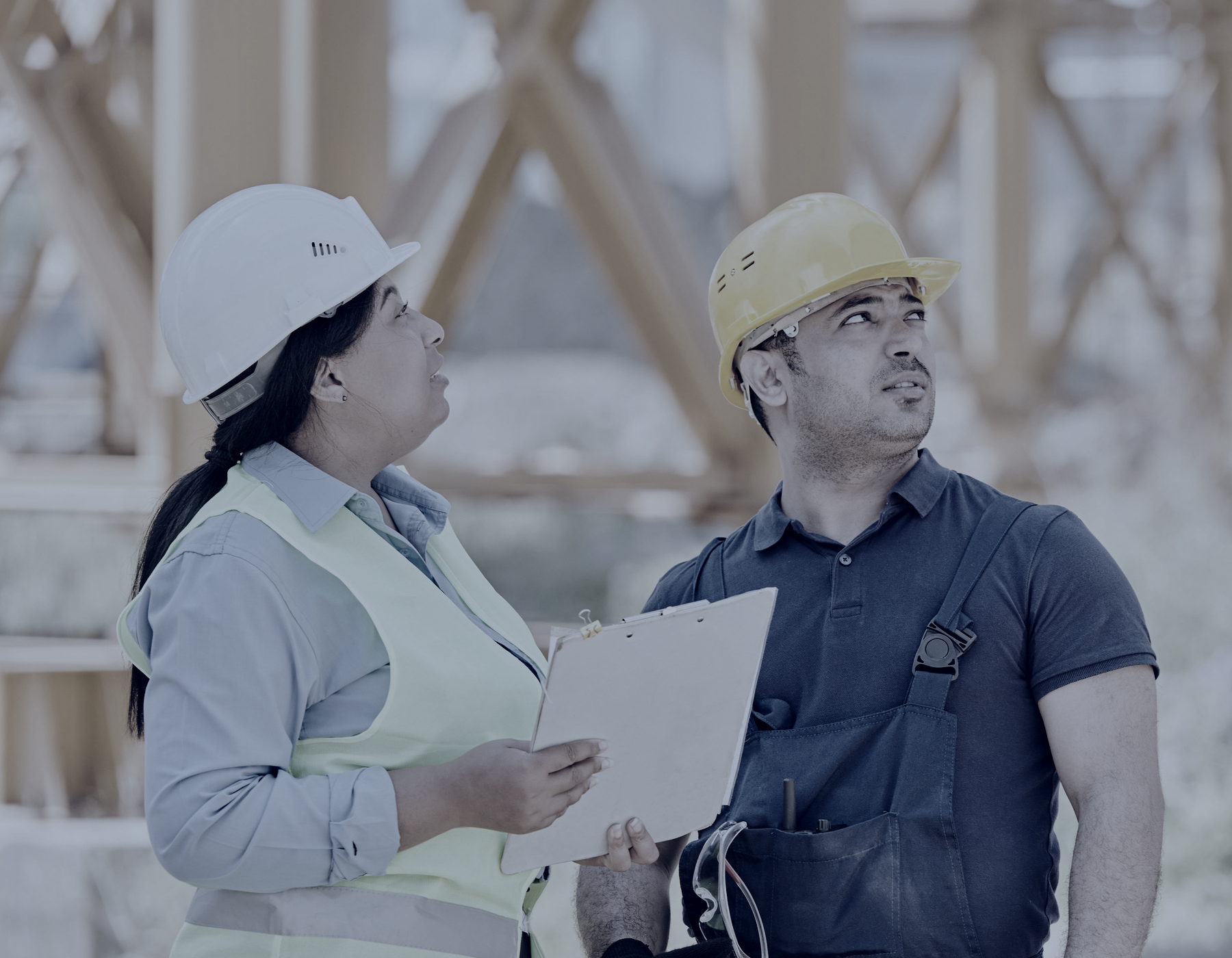 A consultant in a white hard hat and hi-vis vest, clipboard in hand, talking with a site worker in a yellow hard hat and overalls who is looking up at the structure above them.